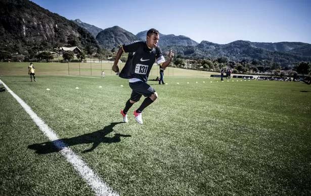 Atleta de roupa preta e dorsal 131 corre em campo de grama, com casas e montanhas ao fundo.