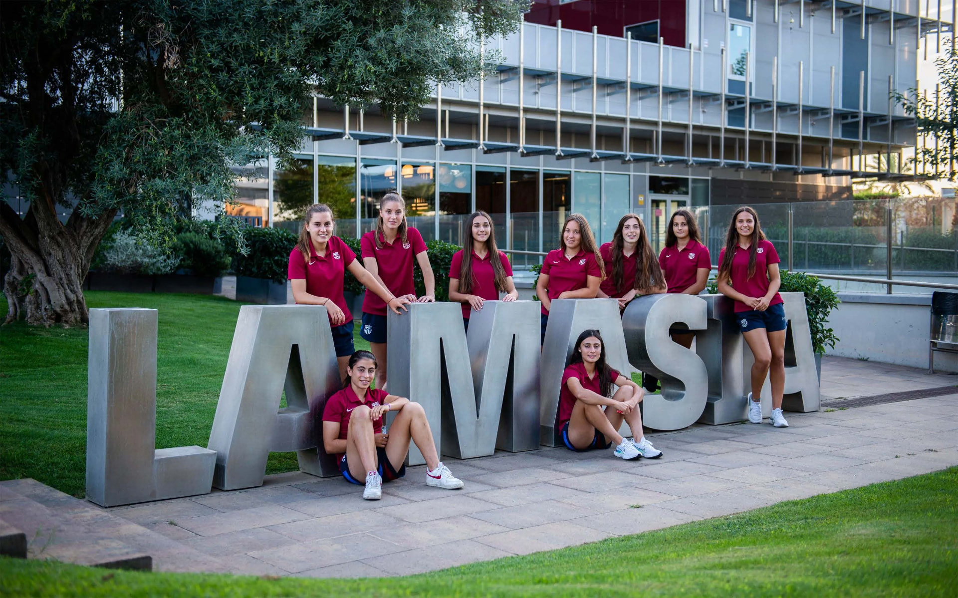 Nove jovens atletas de camisa vinho do FC Barcelona posam sentadas ao lado de letras metálicas que formam a palavra La Masia.