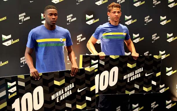 Dois jovens de camisa azul posam diante de um painel do A Chance segurando placas com o número 100 e a marca Nike.