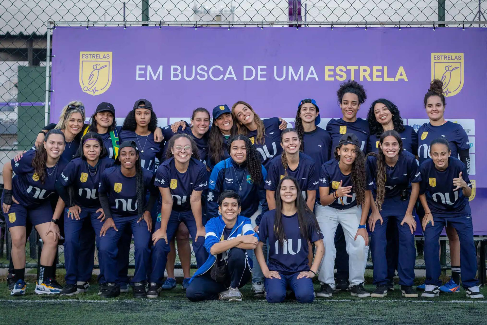 Grupo de atletas e comissão técnica posa em campo sintético diante de faixa roxa com a frase “Em busca de uma estrela”.
